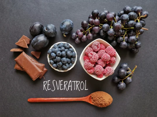 Photo of grapes, plums, blueberries, raspberries, dark chocolate, and a wooden spoon with cocoa powder on a dark grey background. 