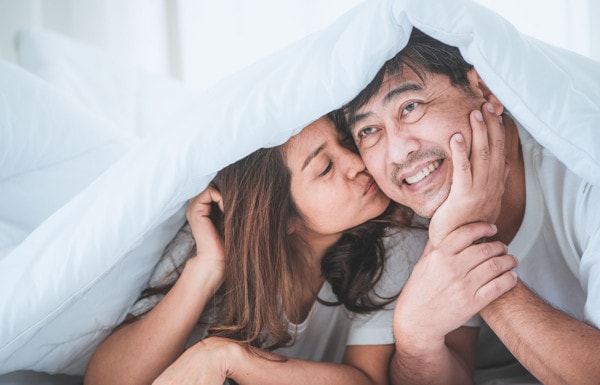 Asian couple in their 40s and 50s playfully under the covers in bed with the woman kissing the man on the cheek