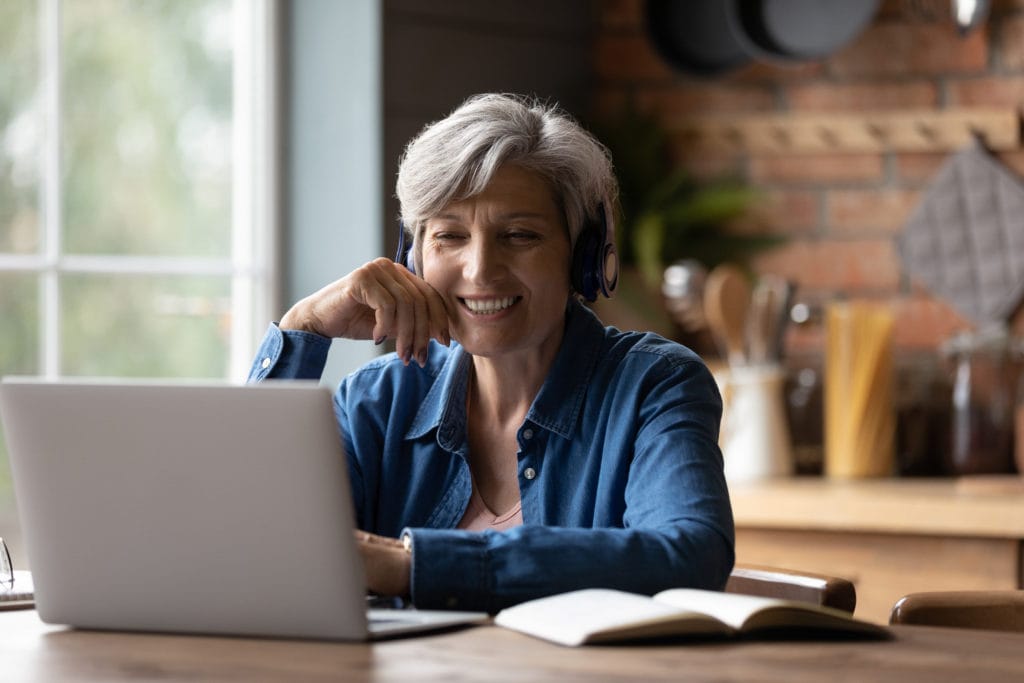 Mature 60s woman in earphones look at laptop screen watch webinar online at home.
