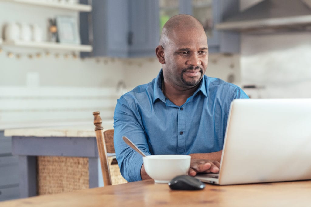 Smiling mature African man sitting at his kitchen table eating breakfast and watching a webinar on his laptop